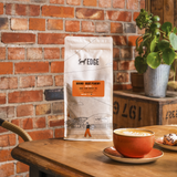 Coffee bag labeled 'EDGE' on a wooden table with a cup of coffee and pastries, against a brick wall.