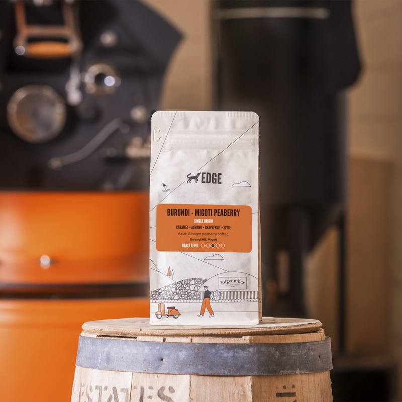 Coffee bag labeled 'EDGE' on a wooden barrel with a blurred coffee machine in the background